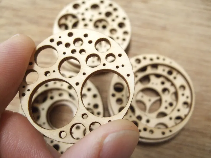 Fingertips holding thin plywood discs cut with patterns of circular holes, with more pieces on the bench