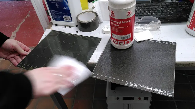 A hand wiping a textured flexible 3D printer build plate with a tissue, next to a bottle of isopropyl alcohol