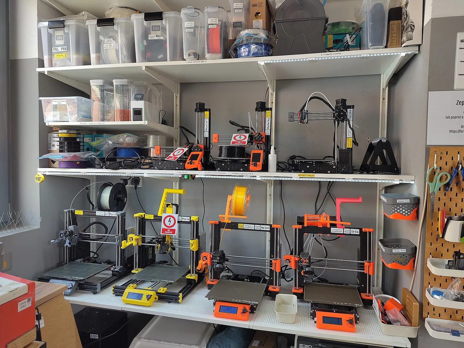 Shelves in a hackerspace holding a row of orange-and-black FDM 3D printers, with filament and parts boxes above