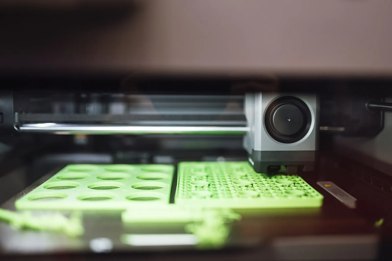 Inside an enclosed 3D printer: the print head moves over a build plate covered in green model parts
