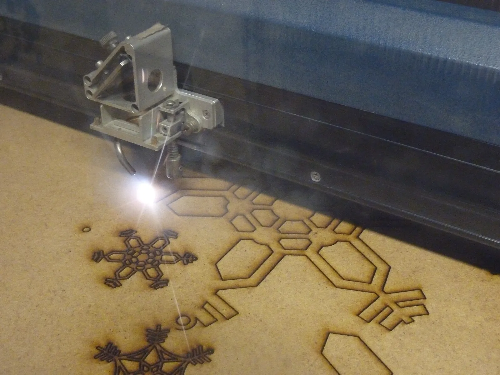 A CO2 laser head cutting snowflake shapes from a sheet of thin wood fibreboard, with a bright point where the beam meets the board